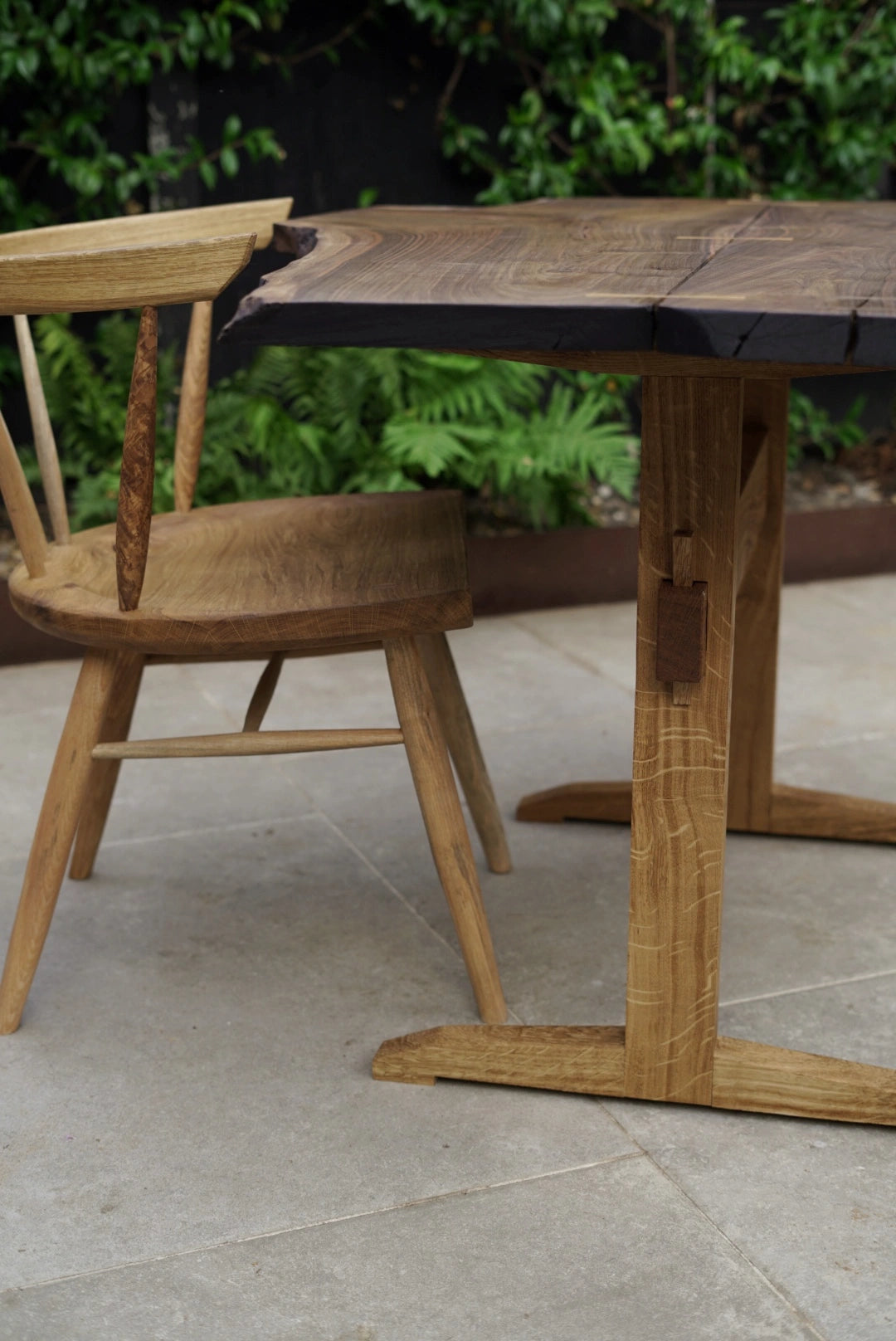 A rustic wooden dining table with a thick live-edge top and a matching wooden chair, set on a light stone patio with green foliage in the background.