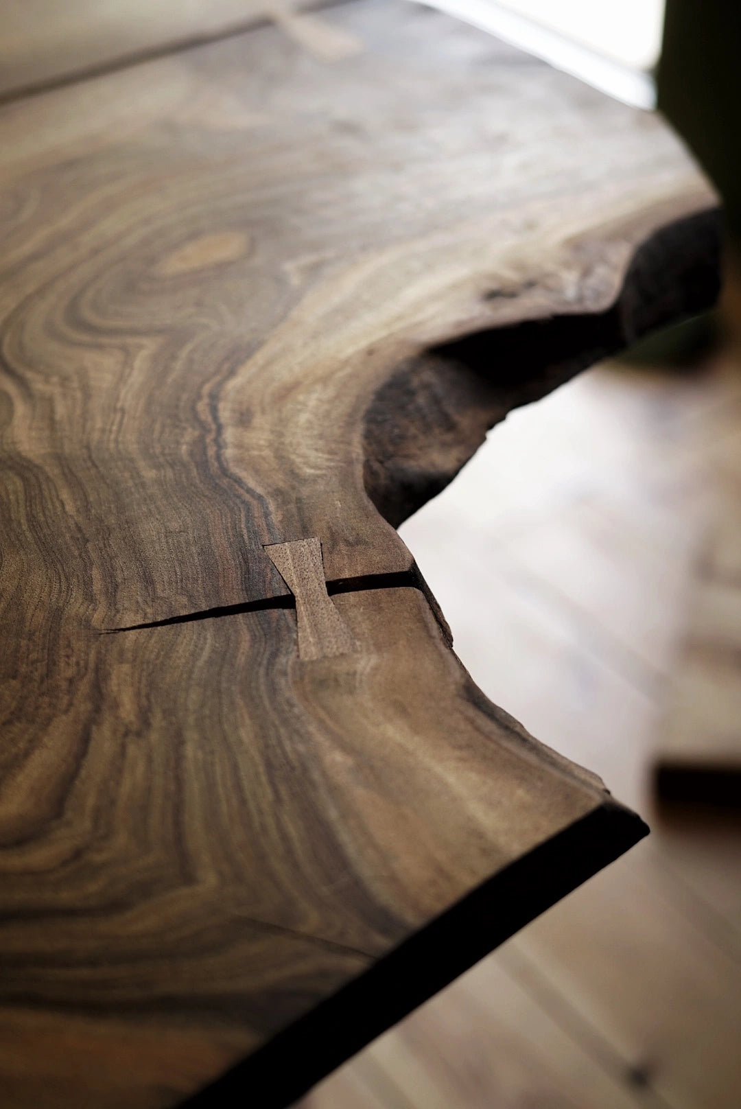 Close-up of a rustic live-edge wooden table showing detailed wood grain patterns and a natural knot, with a blurred warm-toned interior background.