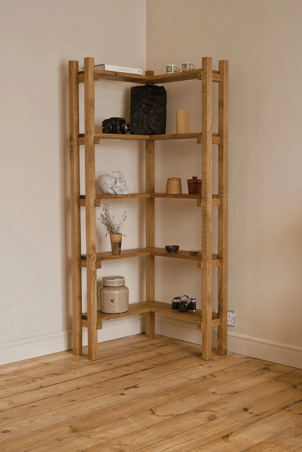 A corner wooden shelving unit made of natural oak, styled with home decor items, set against beige walls and a light wood floor.