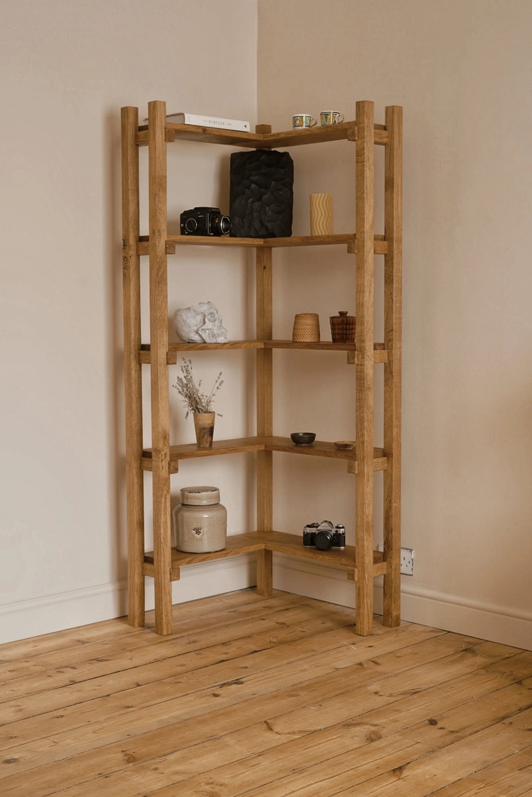 A corner wooden shelving unit made of natural oak, styled with home decor items, set against beige walls and a light wood floor.
