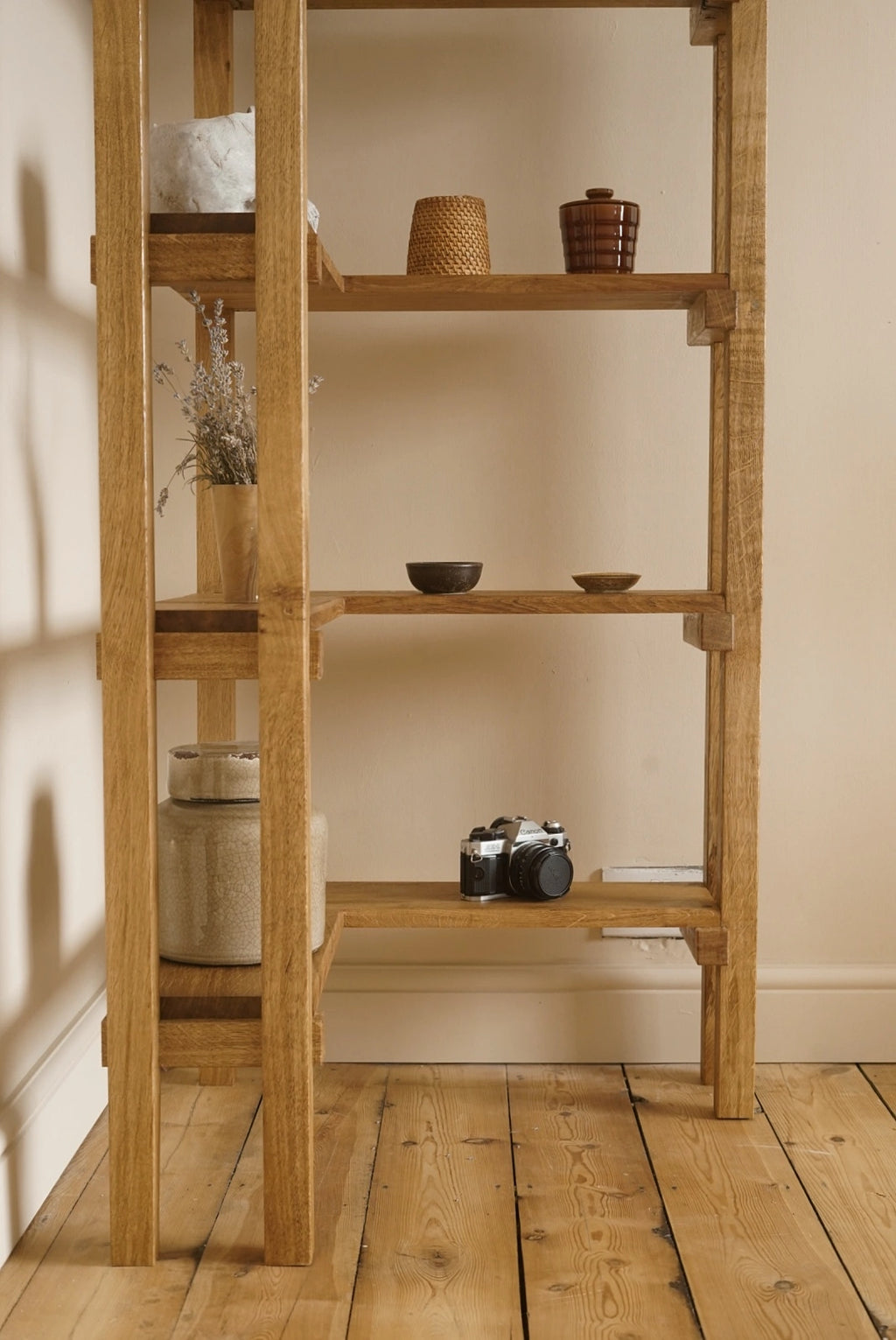 A rustic four-tier wooden shelving unit with a warm natural oak finish, styled with home decor items like ceramic vases, a camera, and dried flowers, set against a beige wall and wooden floor.