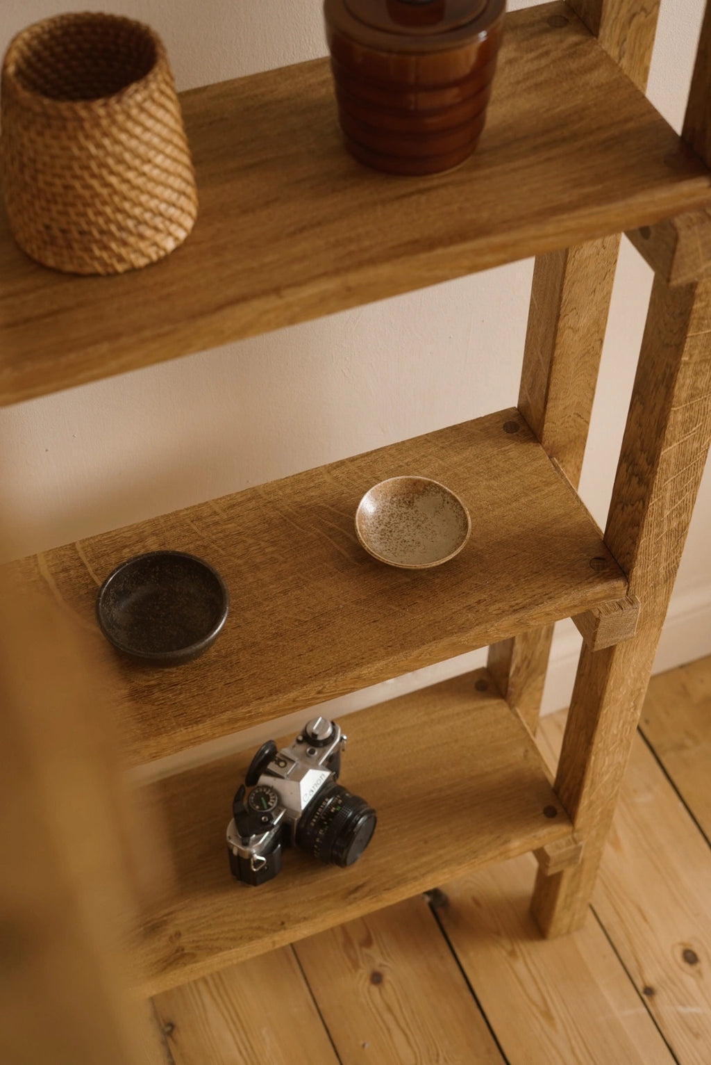 A rustic wooden shelf styled with home decor, including a woven basket, a ceramic jar, a small bowl, a camera, and another dish, set against a neutral wall and light wood floor.