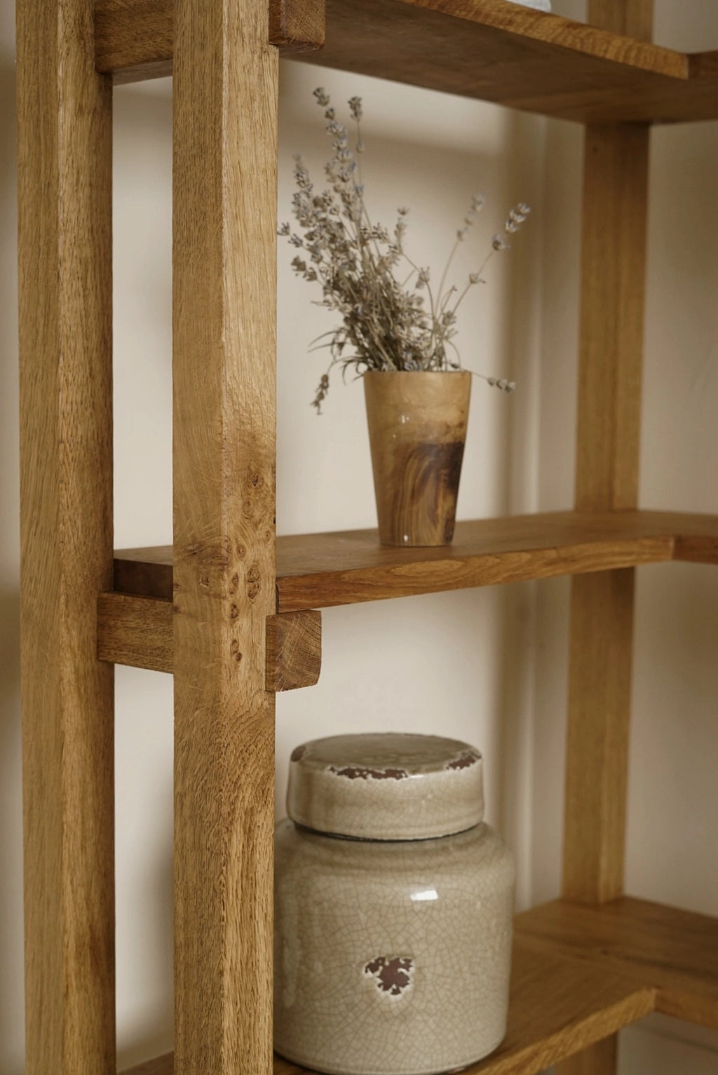 A rustic wooden shelving unit with open shelves, styled with a small ceramic vase holding dried lavender and a large speckled ceramic jar on a lower shelf.